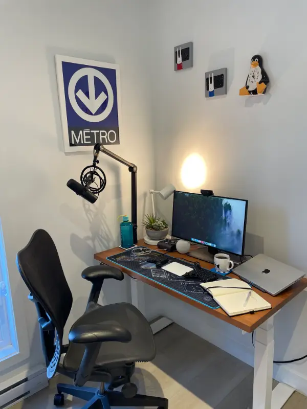 Desk with one monitor, a split keyboard, macbook, and lamp, in a corner of a room with a Montreal Métro poster and Linux clock on the wall above.
