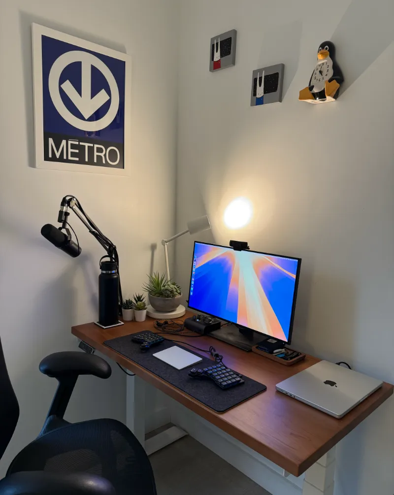 Desk with one monitor, a split keyboard, macbook, and lamp, in a corner of a room with a Montreal Métro poster and Linux clock on the wall above.